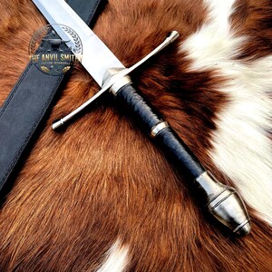 May include: A steel sword with a long, double-edged blade, a straight crossguard, and a black leather-wrapped grip ending in a polished metal pommel. A black leather scabbard lies beside it, featuring a logo with the text THE ANVIL SMITHY.