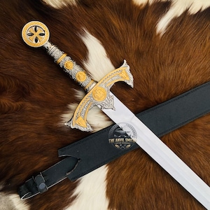 Pode incluir: Uma espada decorativa com punho prateado e dourado com gravações intrincadas e um pomo circular. A guarda tem detalhes dourados e em relevo. Uma bainha de couro preto jaz sob a lâmina, que apresenta o logótipo THE ANVIL SMITH.