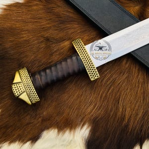 May include: A sword with a dark brown leather-wrapped grip and a brass pommel and crossguard featuring a textured, dotted pattern. The steel blade displays an intricate floral engraving and a circular logo reading The Anvil Smithy.