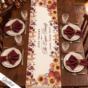 May include: A cream-colored quilted table runner featuring a printed border of sunflowers, pumpkins, and autumn foliage. The center text reads "GIVE THANKS", "The Cooper Family", and "Greatful · Thankful · Blessed" in a dark brown script and serif font.