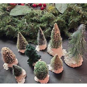 May include: A collection of nine miniature artificial Christmas trees of varying heights and shades of green, each featuring a textured, snow-dusted finish on the branches and a white, uneven base resembling a snowy mound of earth.
