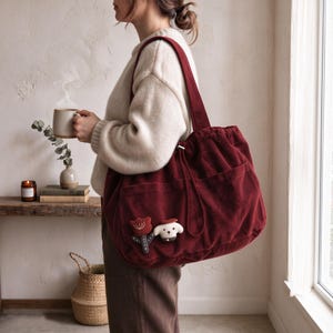 May include: A deep red corduroy tote bag featuring a large main compartment, two exterior pockets, and two decorative plush appliques of a flower and a small dog attached to the front. The bag has long shoulder straps and a drawstring closure.