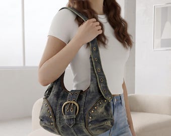 May include: A dark blue, acid-washed denim shoulder bag featuring a large, circular brass-toned buckle on the front flap, decorative brass studs along the sides, and a wide strap with multiple metal grommets for adjustable length.