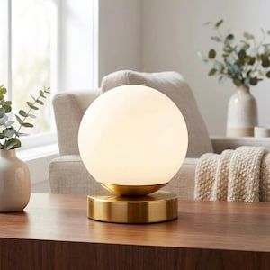 Peut inclure: Une lampe de table composée d'un abat-jour sphérique en verre dépoli blanc reposant sur une base circulaire en laiton brossé. La lampe présente une finition métallique lisse sur le pied et une silhouette arrondie pour le globe en verre.