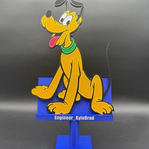 May include: A yellow cartoon dog figure with black ears, a green collar, and a pink tongue, mounted on a blue stand. The stand features a horizontal blue bar with the white text Engineer_KyleBrad printed on the front surface of the base.