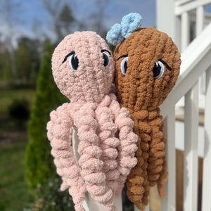 Op de afbeelding: Twee pluche inktvisknuffels gemaakt van zacht, grof garen. De ene is lichtroze en de andere is bruin met een kleine, lichtblauwe gehaakte strik bovenop. Beide hebben grote, geborduurde ogen met zwarte pupillen en witte accenten en gekrulde tentakels.