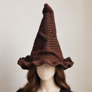 May include: A brown crocheted witch hat with a tall, pointed, and slightly slumped crown. The hat features a wide, wavy brim with a textured, ribbed stitch pattern throughout the entire surface, creating a distinct, layered, and folded appearance.