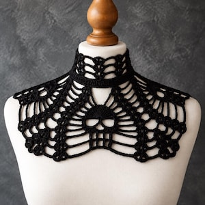 May include: A black crochet collar featuring an intricate web-like pattern with a central skull motif. The accessory is designed to fit around the neck and drape over the upper chest, showcasing detailed needlework and a high-neck choker style.