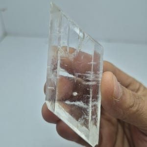 Könnte beinhalten: Ein klares, rautenförmiges Selenit-Kristallexemplar mit sichtbaren inneren Brüchen und Streifen. Das Mineral hat ein durchscheinendes, glasartiges Aussehen mit scharfen, geometrischen Kanten und einer glatten, polierten Oberflächenbeschaffenheit.