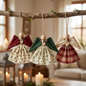 May include: Three hanging angel ornaments made of fabric and wood. The left angel has a dark red wing with gold snowflakes and a cream skirt with holly. The center angel has green wings with white dots and a cream skirt with mistletoe. The right angel has cream wings with gold snowflakes and a red plaid skirt. Each has a wooden bead head, a small heart charm, and a twine loop.