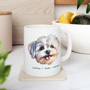 May include: A white ceramic mug featuring a colorful illustration of a Maltese dog's face with its tongue slightly out. Below the dog, the text reads Coffee > Code > Maltese in a dark teal font. The mug has a standard handle and a smooth finish.