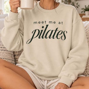 May include: A beige crewneck sweatshirt featuring the black text "meet me at pilates" printed on the front. The garment has long sleeves with ribbed cuffs and a ribbed neckline, constructed from a solid-colored fabric without patterns.