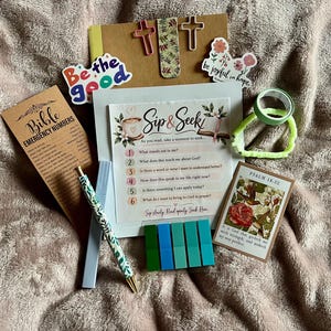 May include: A devotional kit featuring a Sip & Seek card, a Bible Emergency Numbers bookmark, a floral pen, a Psalm 18:32 card, cross-shaped paperclips, stickers reading Be the Good and be joyful in hope, a light green tape roll, and five colored tabs.