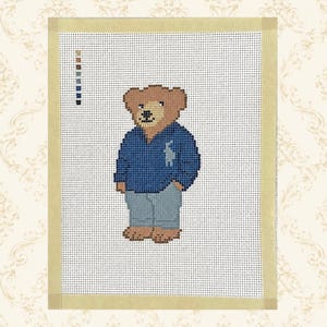 May include: A needlepoint canvas featuring a cross-stitched brown bear wearing a blue long-sleeved shirt and light gray pants. The bear has a small logo on the chest of the shirt. A vertical color key is visible on the left side of the canvas.