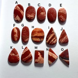 May include: A collection of fifteen polished red and cream-colored jasper cabochons arranged in three rows, labeled A through O. The stones feature various shapes including ovals, circles, teardrops, rectangles, and triangles with distinct patterns.