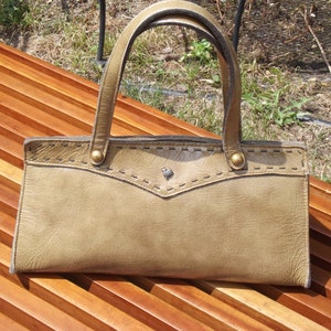May include: A tan leather handbag with a stitched design and two gold-toned clasps. The bag is sitting on a wooden surface.