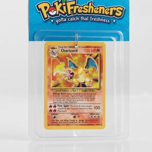 May include: A PokiFresheners air freshener in retail packaging featuring a Charizard trading card design. The card is yellow and orange with a fire-breathing dragon illustration, 120 HP, and text including Fire Spin. The header card is blue.
