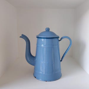 May include: A light blue enamel coffee pot with a matching lid, a curved spout, and a side handle. The cylindrical body features a smooth, glossy finish with subtle signs of wear along the base rim and the spout tip, showcasing its construction.