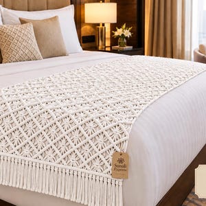 May include: A cream-colored macrame bed runner featuring an intricate diamond-shaped knot pattern and long fringe trim at the ends. The runner is draped across a white bedspread and includes a small brown paper tag labeled Sunali Exports.