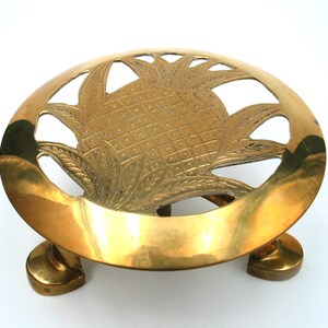 May include: A brass trivet with a pineapple design. The trivet is oval-shaped and has a cutout pineapple in the center. It stands on three rounded feet and is made of shiny, polished brass.