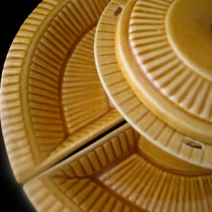 May include: A three-sectioned, tan-colored serving dish with a lid. The dish and lid have a radial, ridged design. The lid has a dark brown handle. The dish is likely made of ceramic or a similar material. The background is black.