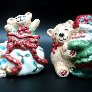 May include: Two ceramic Christmas figurines. One is a teddy bear emerging from a gift bag with a red bow and holly accents. The other is a teddy bear hugging Santa Claus. Both figurines have smiling faces and are painted in festive colors.