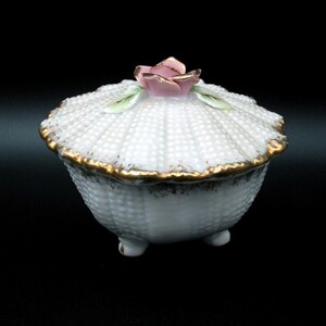 May include: A white porcelain trinket box with a textured, dotted pattern. The lid is topped with a pink rose and green leaves. The rim is trimmed with gold. The box sits on three small feet.