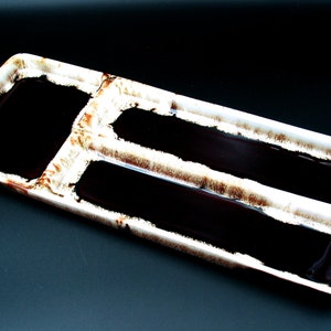 May include: A rectangular ceramic serving dish with three compartments and a white and brown rim. The compartments are filled with a dark, glossy substance, possibly a sauce or glaze. The dish is set against a dark background.