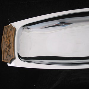 May include: A long, rectangular serving tray with a polished, reflective silver-tone metal surface. The tray features a decorative, dark brown, textured handle on one end with a carved, wing-like motif and two circular holes.