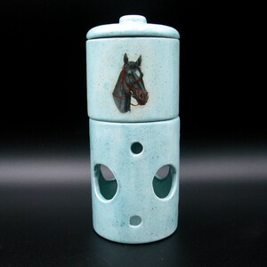 May include: A two-piece ceramic container in a light blue hue. The top section features a lid and a painted horse head with a red bridle. The bottom section has three circular cutouts. The container is approximately 6 inches tall.