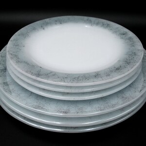 May include: A stack of five white and light blue plates. The plates have a white center and a light blue rim with a dark gray, abstract pattern. The plates are stacked on a black background.