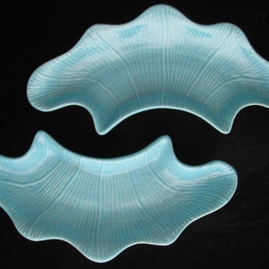 May include: Two light blue, uniquely shaped ceramic dishes. Each dish has a wavy, organic form with a textured, fan-like pattern in white. The dishes are set against a black background, highlighting their color and design.
