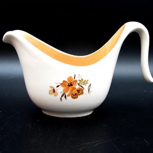 May include: White ceramic gravy boat with a curved handle and a floral design in shades of brown and yellow. The rim is accented with a band of orange. A classic serving dish for sauces and gravies.