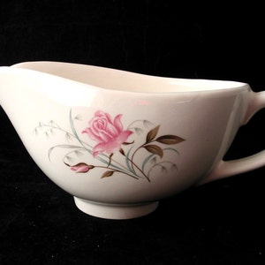 May include: White ceramic gravy boat with a pink rose and floral design. The gravy boat has a spout, handle, and a decorative rose and floral pattern. The design includes pink roses, brown leaves, and light blue accents.