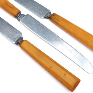 May include: Four butter knives with stainless steel blades and amber-colored handles are arranged diagonally on a white background. The handles have a rectangular shape with a slight taper, and the blades are slightly curved.