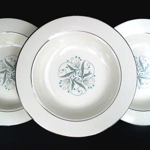 May include: Three white ceramic soup bowls featuring a central floral motif with teal leaves and thin gray swirling lines. Each bowl has a smooth, wide rim accented with a thin silver metallic band along the outer edge, creating a clean look.