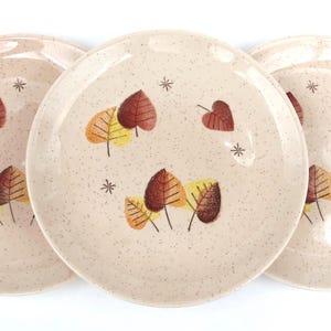 May include: Three beige ceramic plates with a speckled finish. Each plate features a design of stylized autumn leaves in shades of brown, yellow, and orange, along with small star accents. A set of decorative tableware.