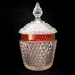 May include: A clear glass jar with a lid. The jar has a diamond pattern and a red band around the middle. The lid is clear and has a pointed finial. The jar is on a black background.