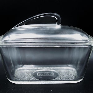 May include: A clear glass rectangular butter dish with a matching lid featuring a curved handle. The base of the dish has a textured bottom surface and a small embossed rectangular logo in the center. The glass is transparent and smooth.