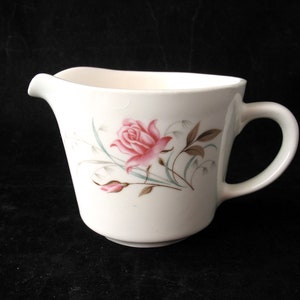 May include: White ceramic creamer with a pink rose floral design. The creamer has a curved spout and a handle. The rose is surrounded by green and brown leaves and stems. The creamer is on a black surface.