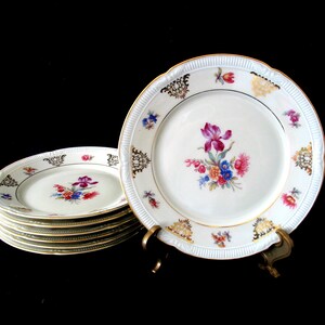 May include: A stack of white porcelain plates with scalloped edges and gold trim. Each plate features a central floral bouquet in shades of pink, purple, blue, and orange, surrounded by smaller floral accents and ornate gold filigree patterns.