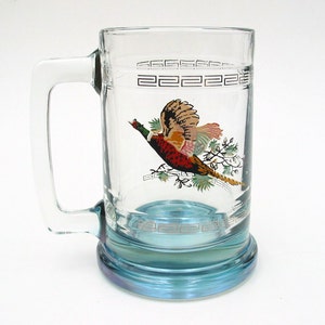May include: Clear glass beer mug with a blue base and handle. The mug features a colorful illustration of a pheasant in flight, with a decorative border near the top. A classic design for serving beer or other beverages.