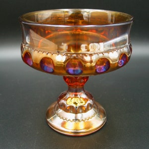 May include: An iridescent orange glass pedestal bowl. The bowl has a wide, shallow top and a decorative band of circles with purple and blue accents. The stem and base are also orange, creating a cohesive design.