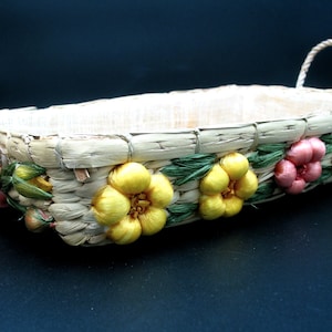 May include: A rectangular woven basket with two rope handles. The basket is decorated with yellow and pink fabric flowers and green leaves along the sides. The basket is made of natural-colored straw.