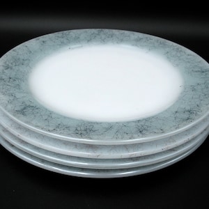 May include: A stack of four white plates with a light blue and black abstract design around the rim. The plates have a glossy finish and are stacked on a black surface.