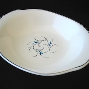 May include: Off-white oval serving bowl with a black rim and a silver accent. The bowl features a delicate teal and black floral design in the center. The rim has a scalloped edge, adding a touch of elegance to the vintage tableware.