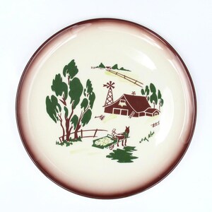 May include: A cream-colored ceramic plate with a brown rim and a farm scene design. The scene includes a red barn, green trees, a windmill, and a horse-drawn cart. The plate is round.