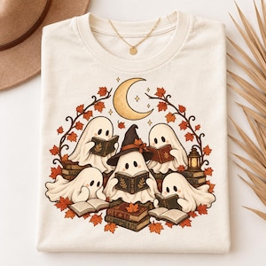 May include: A cream-coloured t-shirt featuring a graphic illustration of five white ghosts wearing a witch hat and reading books. The design includes a crescent moon, autumn maple leaves, and stacks of books arranged in a circular pattern.