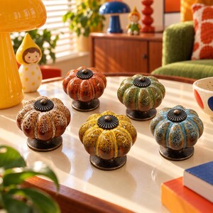 May include: Five ceramic cabinet knobs shaped like pumpkins, each featuring a unique glazed finish in shades of orange, green, brown, yellow, and blue. Each knob has a dark bronze-colored metal base and a matching circular top accent.
