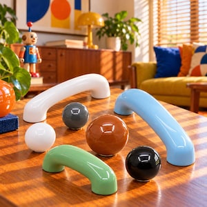 May include: A collection of cabinet hardware including two arched handles in white and light blue, and four spherical knobs in white, dark gray, brown, and black. Each piece features a smooth, glossy finish and a solid, rounded design.